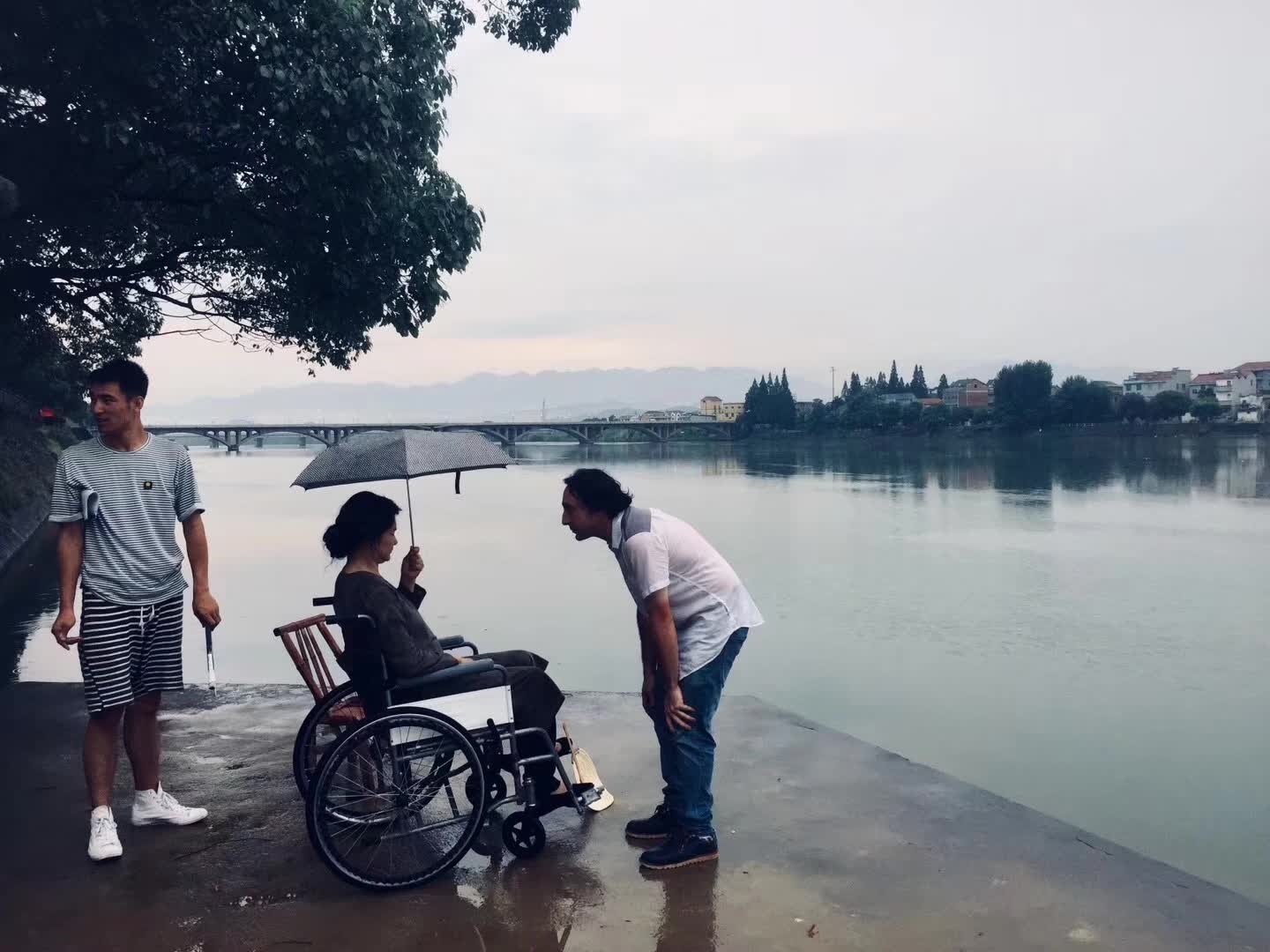 Three people stand beside a river, including a person seated in a wheelchair under an umbrella.